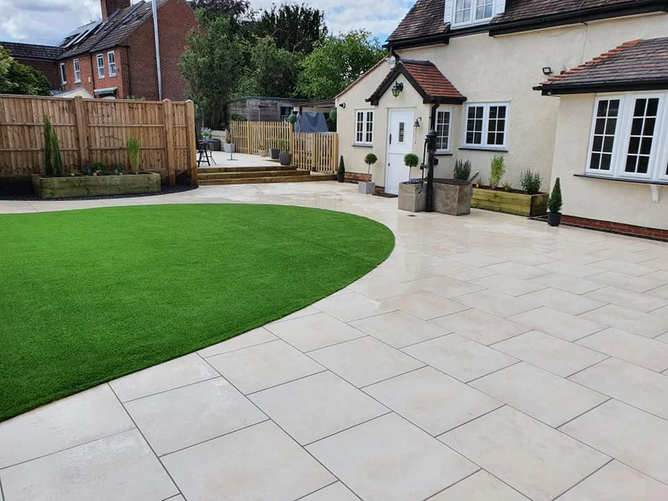 A spacious backyard with a large area of beige paving stones, a patch of green artificial grass, and a white house with multiple windows. There are planters with small trees and potted plants, a wooden fence, and steps leading to a raised deck.