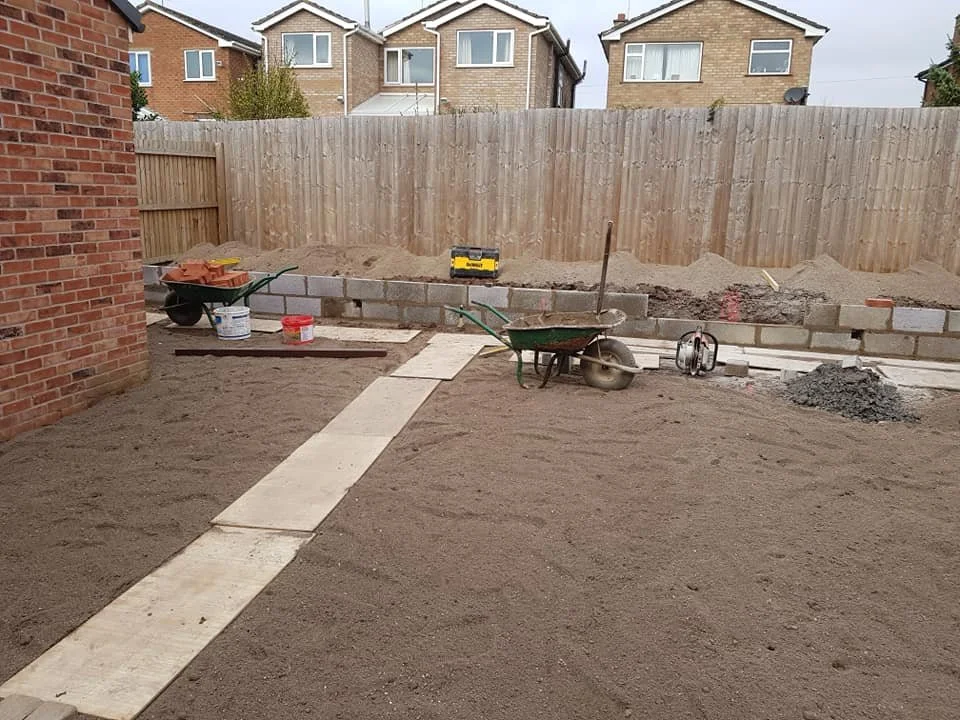 Backyard under construction with a pathway made of concrete slabs, construction tools, sand piles, and a wooden fence.