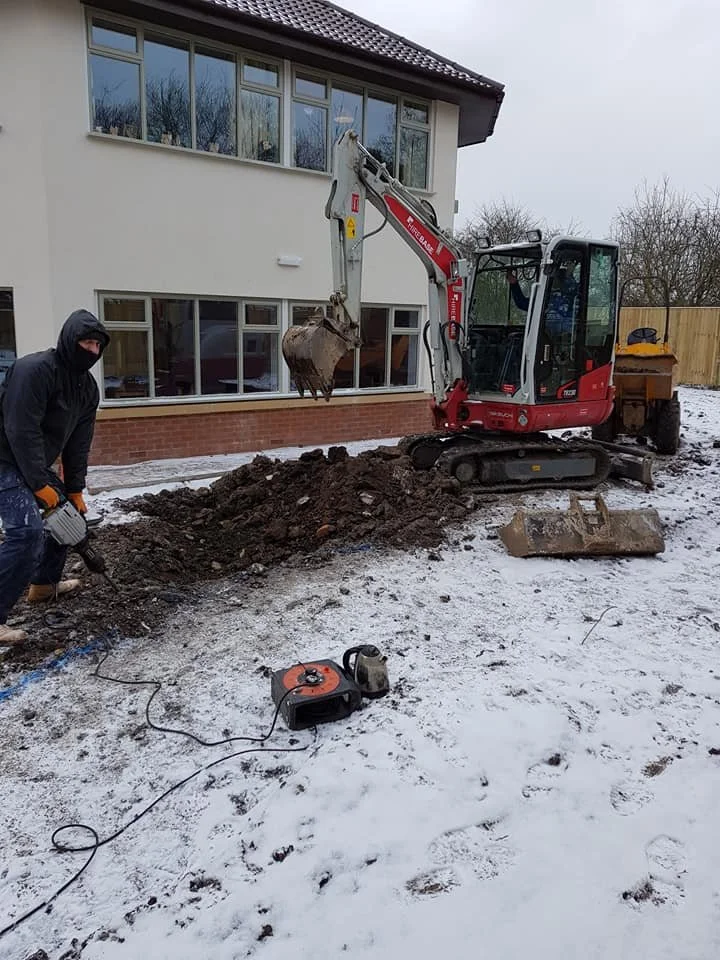 A construction worker in a black hoodie and orange gloves using a jackhammer on a dirt patch outside a white house with large windows. A small red and black excavator is parked nearby, and there is snow on the ground.