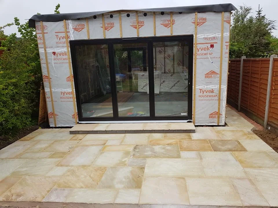 Small building with sliding glass door, partial construction with Tyvek Housewrap, and a patio with large tan tiles.