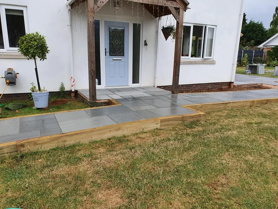 Newly constructed front porch with gray paving stones, wooden border, and a small tree in a pot, attached to a white house with a blue front door and a wooden canopy supported by beams.