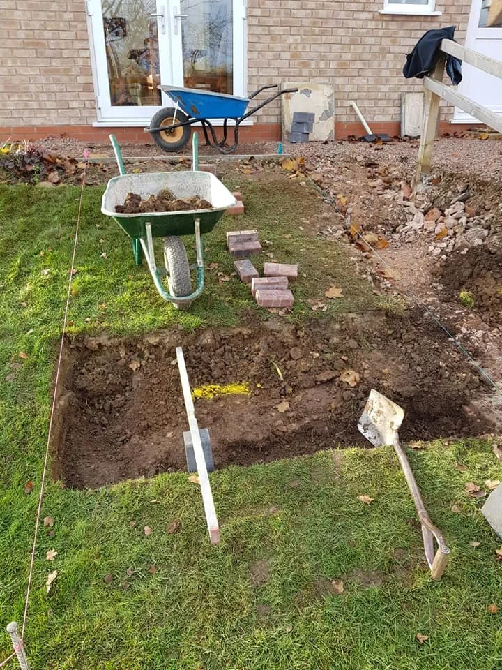 Garden trench excavation with a wheelbarrow, bricks, and gardening tools near a house with a brick wall and a glass door.