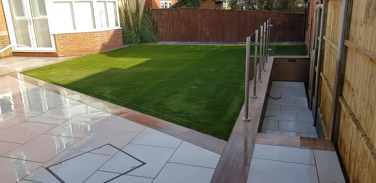 Backyard with a small patch of vibrant green artificial grass, a patio with shiny large white tiles, a wooden fence, and a building with a brick exterior and large windows.