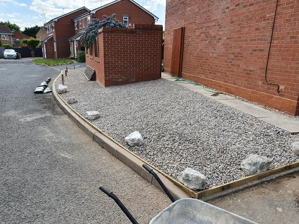 Construction site on a residential street with gravel filling and wooden edging, a brick wall and house, construction tools, and large rocks along the edge.