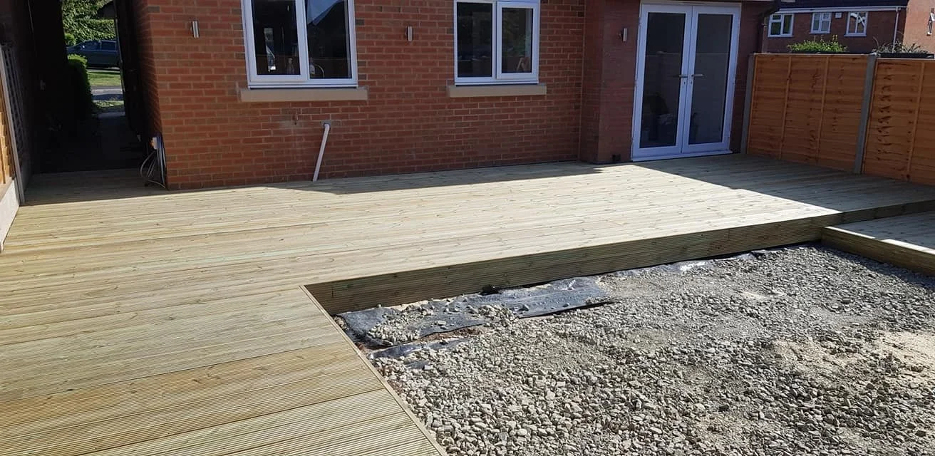 A newly constructed wooden deck outside a brick house, with a section of gravel and dirt showing unfinished ground.