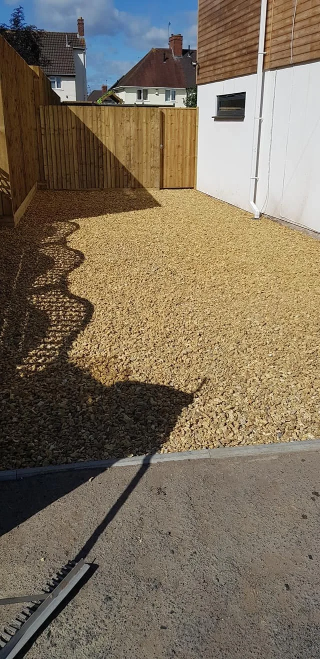 A backyard with a gravel ground, wooden privacy fence, and a white building wall with a small window. Shadows of a rake and a person are cast on the gravel, and neighboring houses with rooftops are visible in the background.