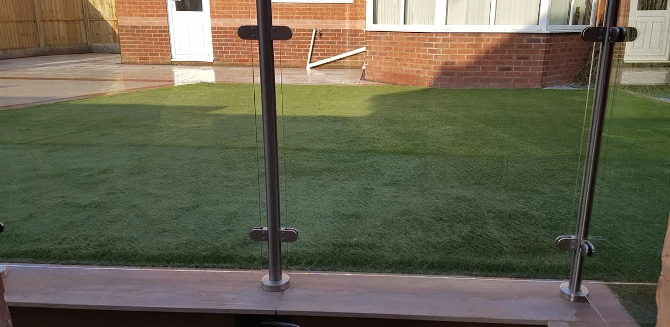 Backyard with artificial grass, brick house, glass fence, wooden deck, and patio area.