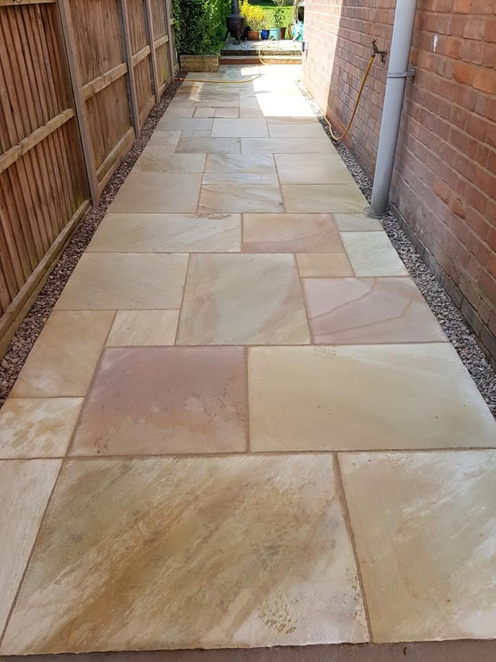 A newly paved patio with beige and light brown stone tiles, bordered by a wooden fence on the left and a brick wall on the right, leading up to a garden area with potted plants and steps.