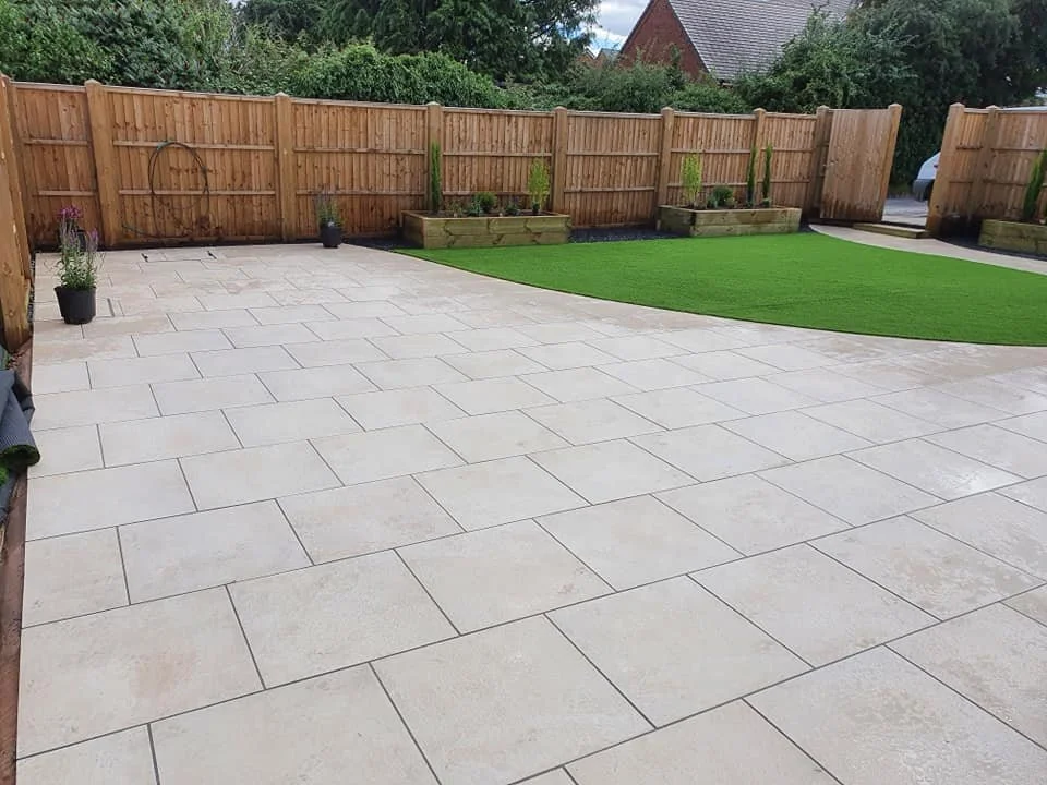A backyard patio with large light-colored paving stones, a wooden fence, potted plants, and a patch of green artificial turf surrounded by garden beds with plants.