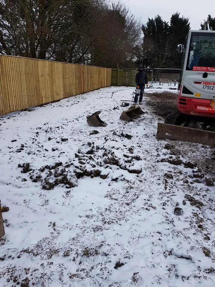 Part of a construction site covered in snow with a bulldozer blade on the ground. A worker wearing cold-weather clothing is standing near the fence. A large construction vehicle is parked to the right.