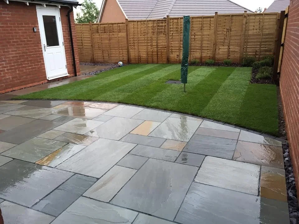 Backyard with a wet stone patio and a well-maintained grassy lawn enclosed by a wooden fence, with a soccer ball near the fence and a closed umbrella in the middle of the lawn.