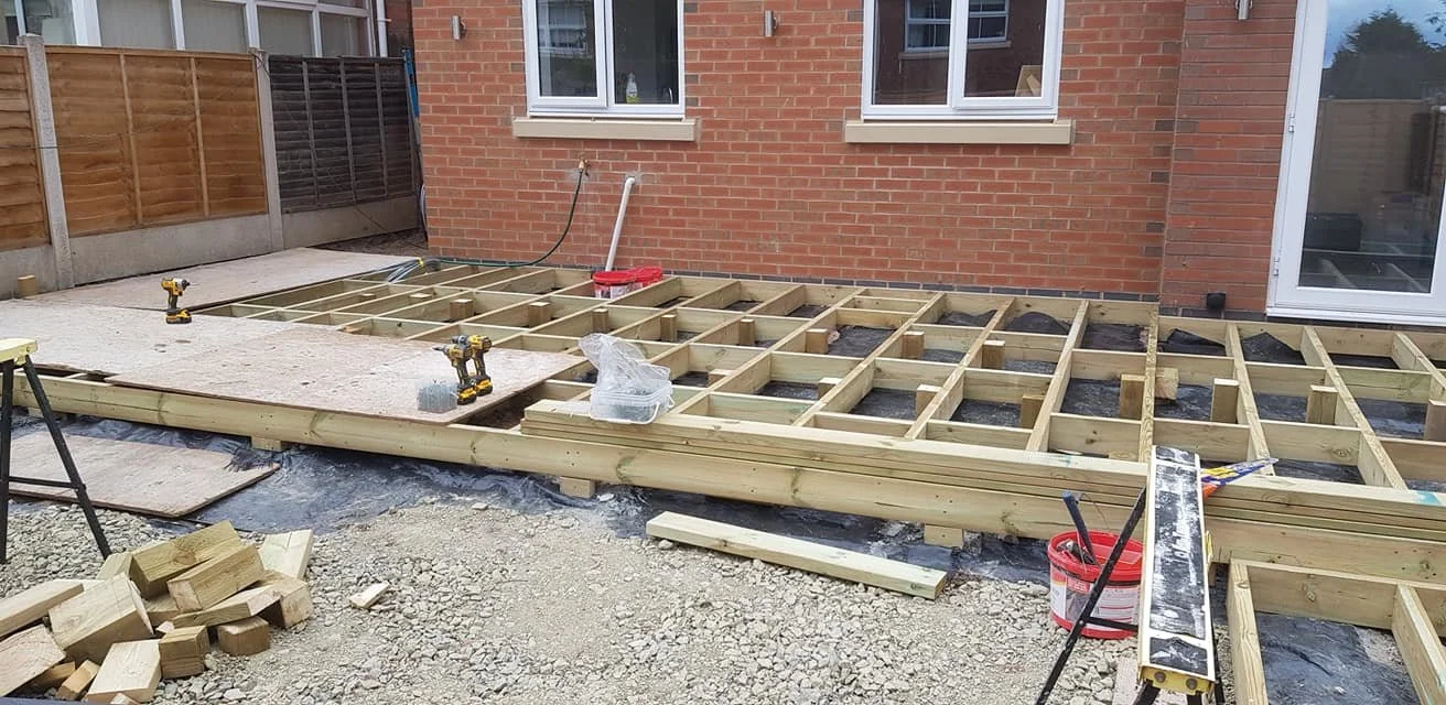 Construction site showing wooden framework for a deck or patio being built outside a brick house, with various tools and materials scattered around.