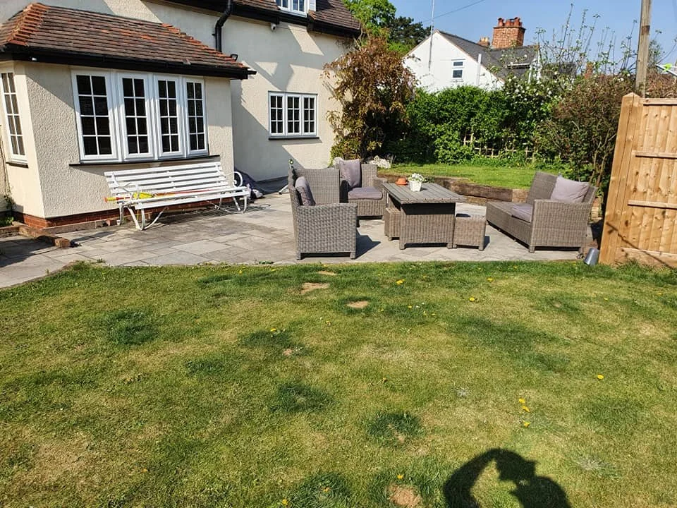 A backyard patio with outdoor furniture, including a set of four gray wicker chairs with cushions and a matching coffee table, surrounded by a lawn and a wooden fence. There is a white house with multiple windows and a bench on the patio.