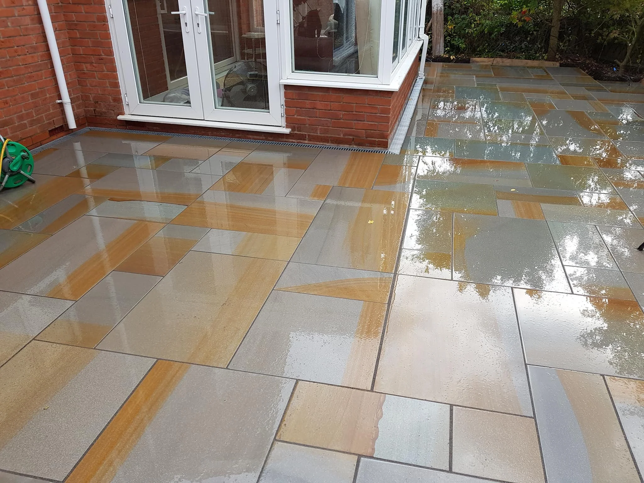 Wet, freshly cleaned outdoor patio with multicolored tiles, next to a brick house with a glass door, and a garden with trees and shrubs.