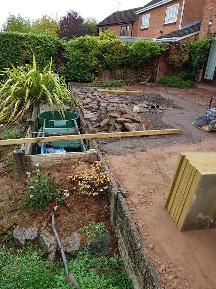 Backyard garden undergoing renovation with paving stones, construction tools, and materials visible, including a green water barrel, wooden planks, and a yellow building block.