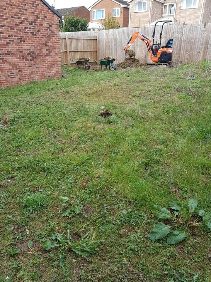 Backyard with a small orange and black excavator digging near a fence, two green wheelbarrows, and a brick house wall on the left.
