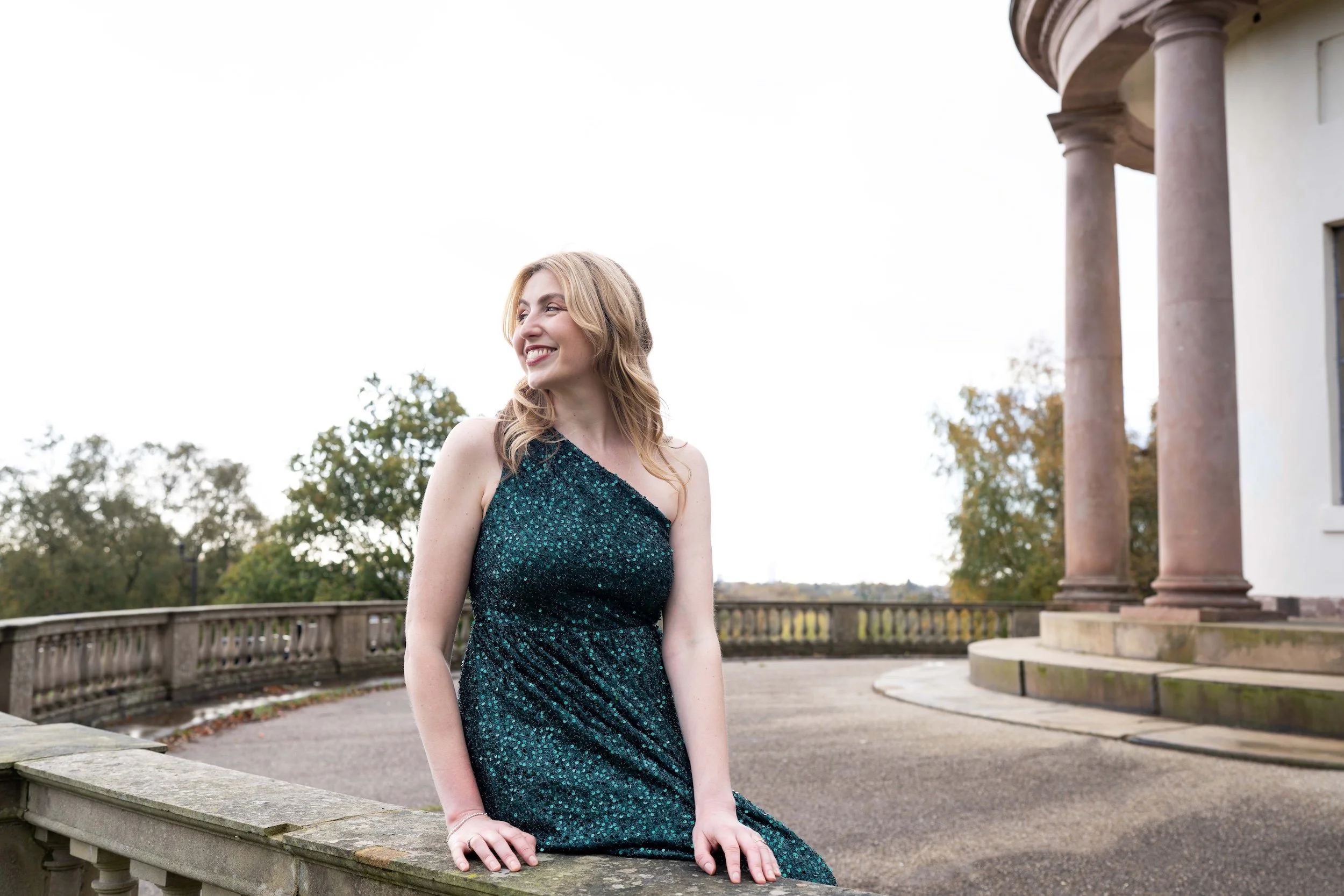 A woman in a sparkly green dress sitting outdoors on a stone ledge, smiling and looking to her left, with a building with large columns and trees in the background.