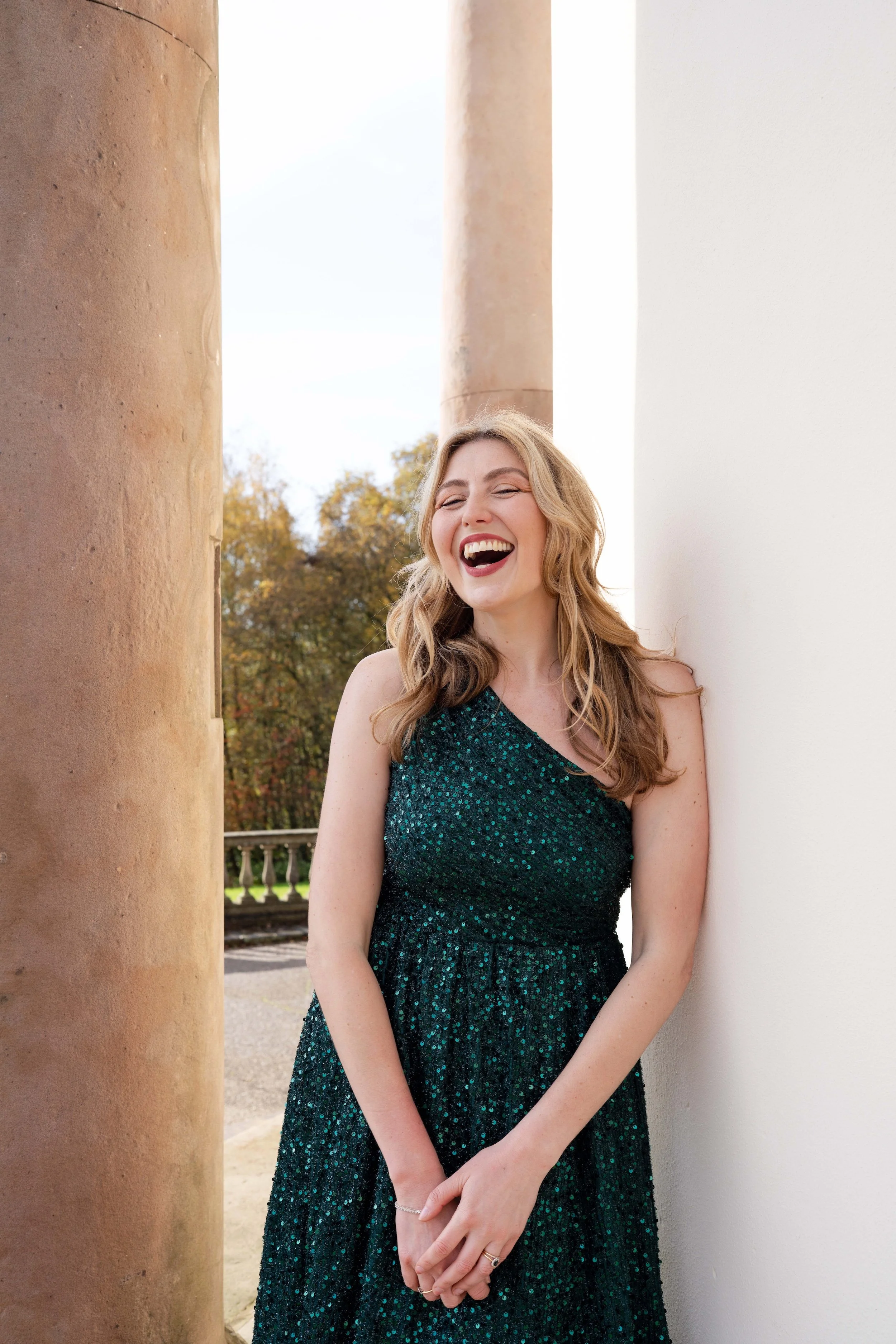 A woman with long wavy blonde hair is standing outdoors, leaning against a white wall and laughing happily. She is wearing a one-shoulder, sparkling dark green dress, with trees and columns visible in the background.