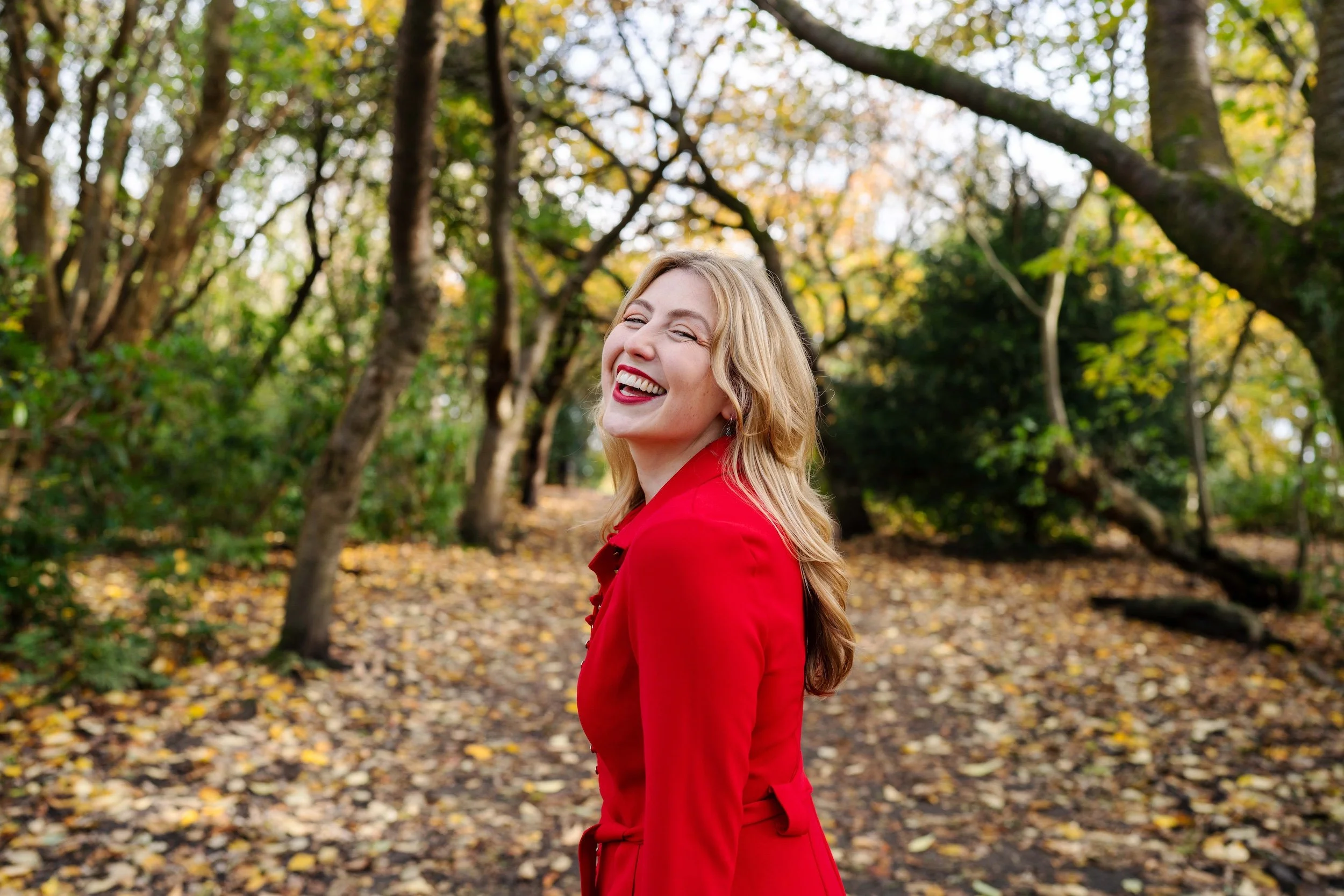 A woman with blonde hair, wearing a red coat, smiling and laughing in a forest with autumn leaves on the ground.