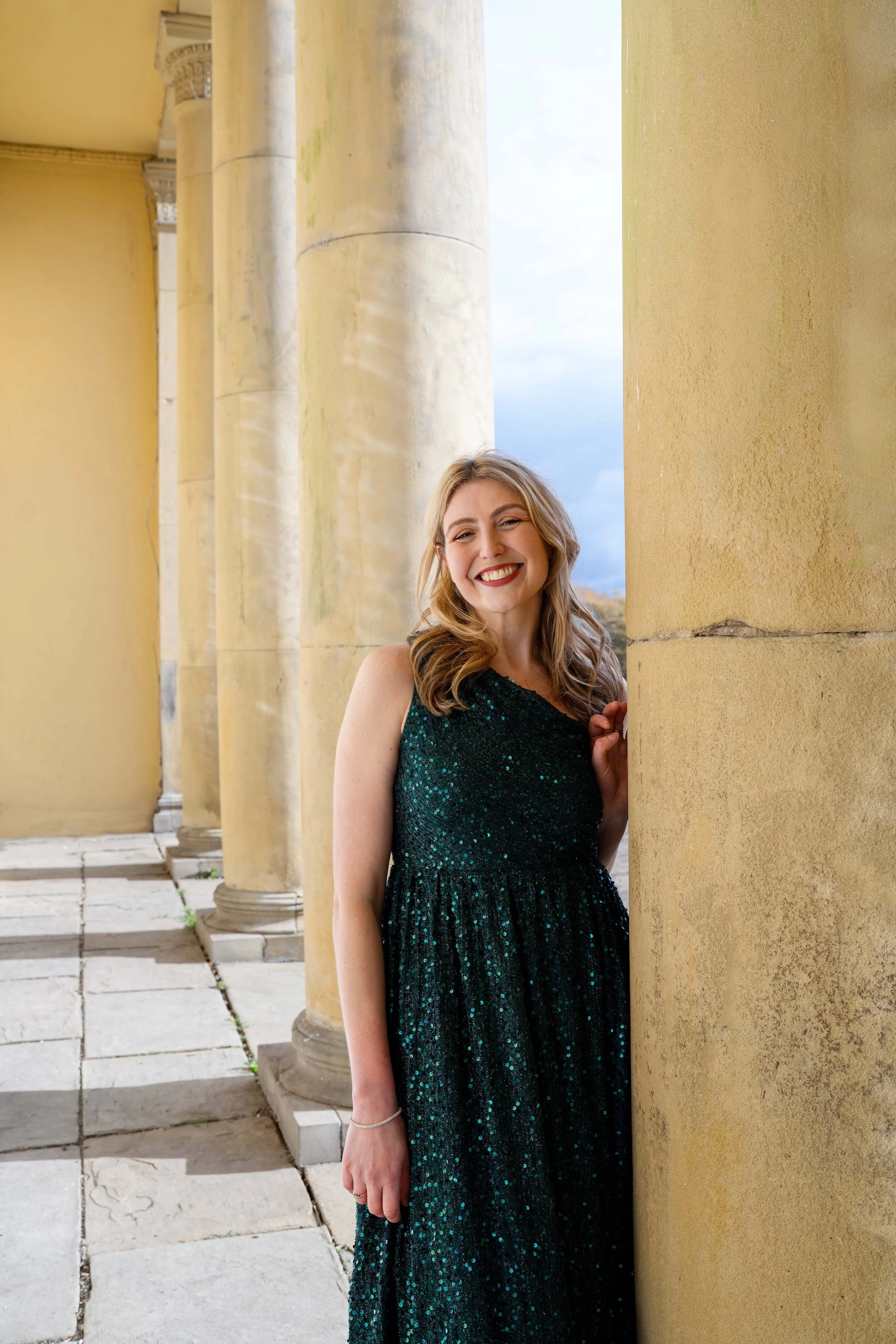 A woman with long blonde hair and a big smile, wearing a dark green sequined dress, stands beside a large stone column on a sunny day.