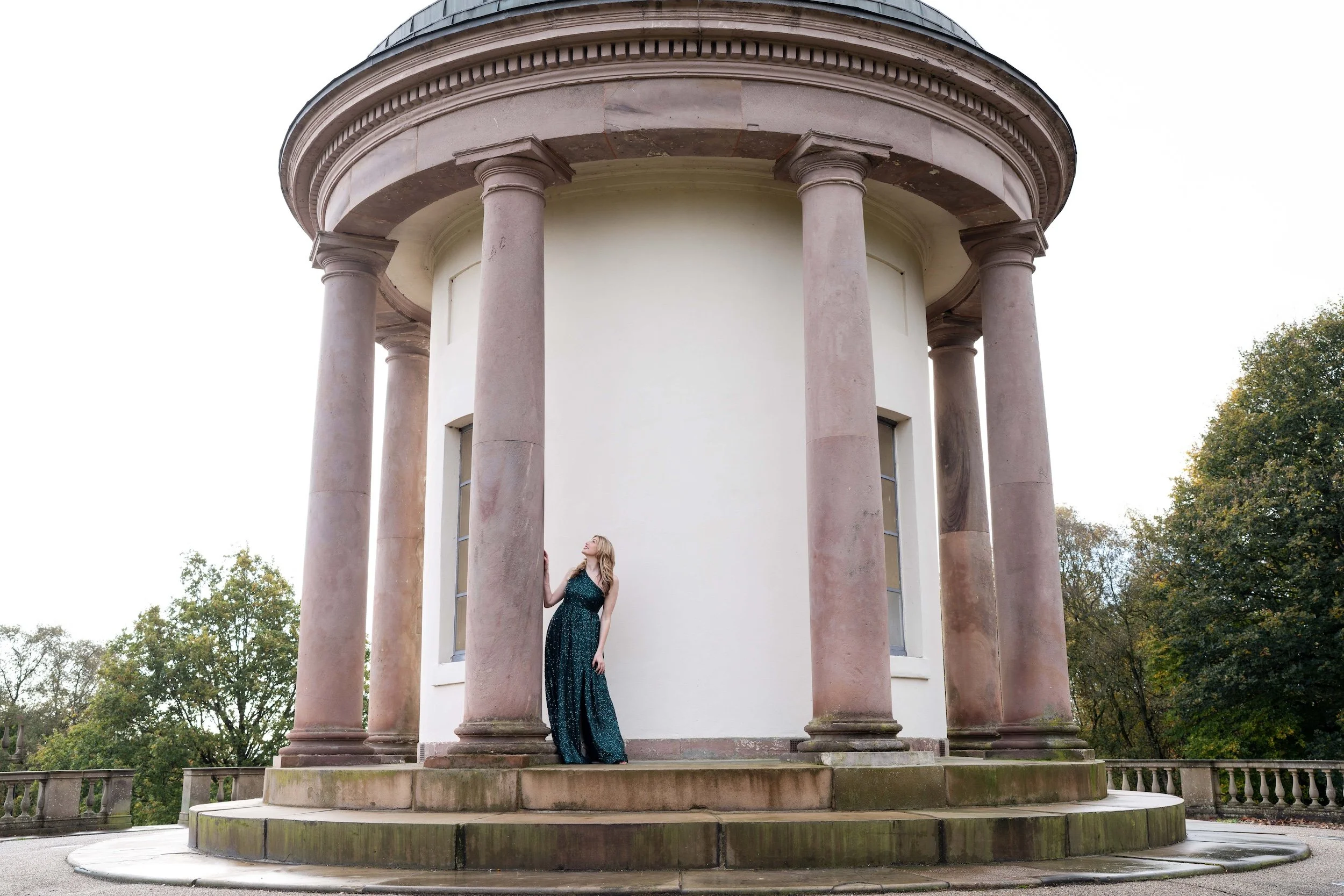 A woman in a long, dark green dress stands on the steps of a circular, white building with tall, pinkish columns and a dome, surrounded by trees.