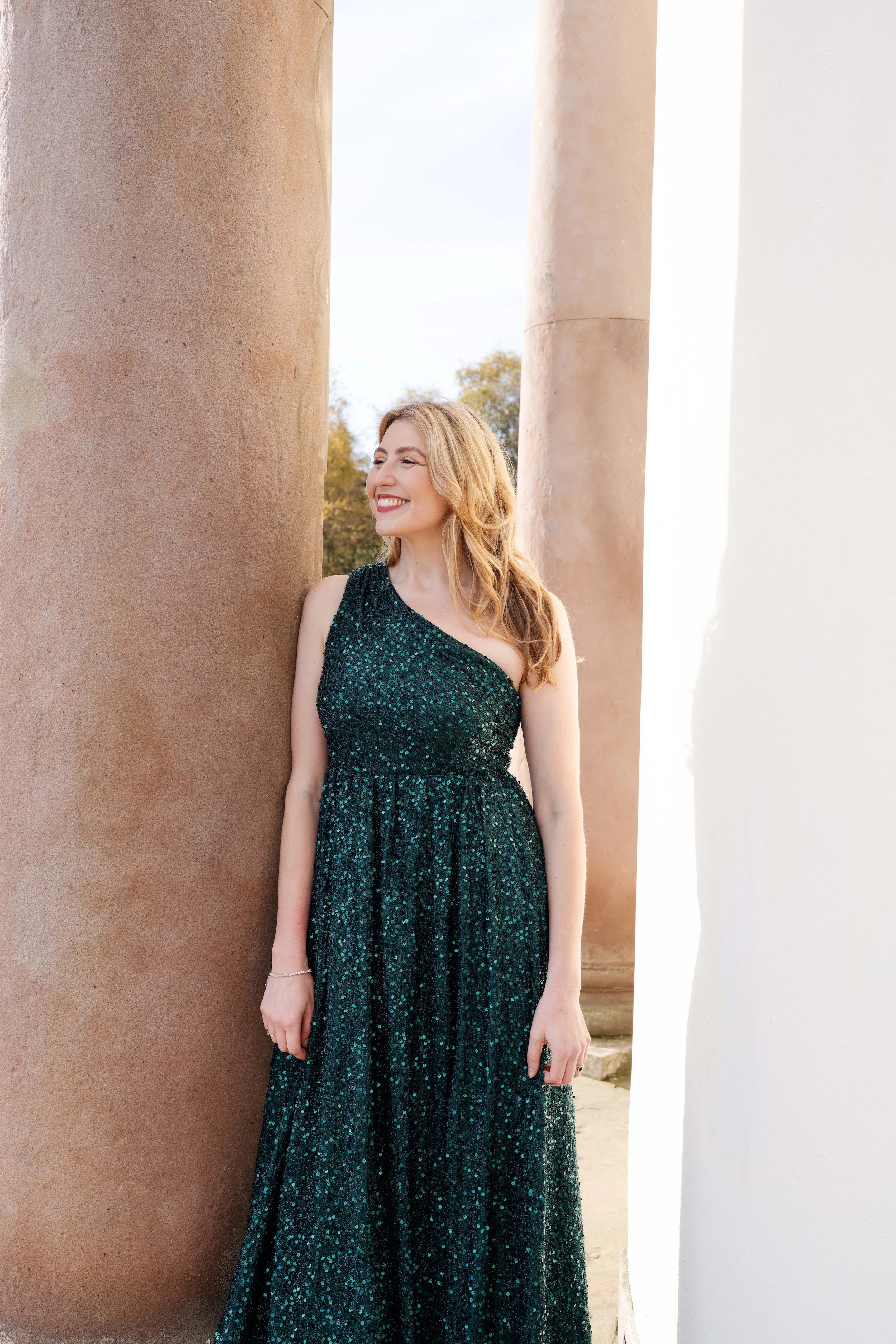 A woman in a sparkly, dark green dress with one shoulder, standing between two columns outdoors and smiling.