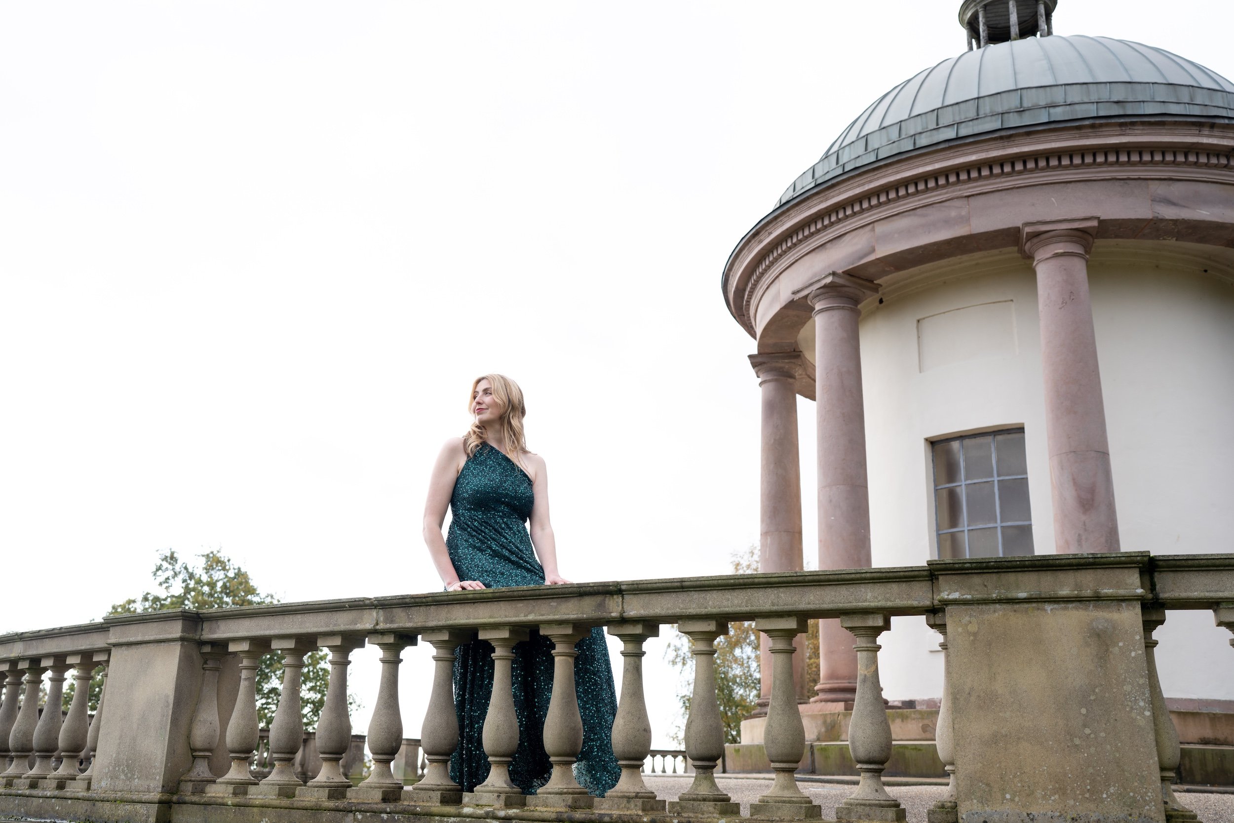A woman in a sparkly dark green dress standing on a stone balcony with a classical building and dome in the background.