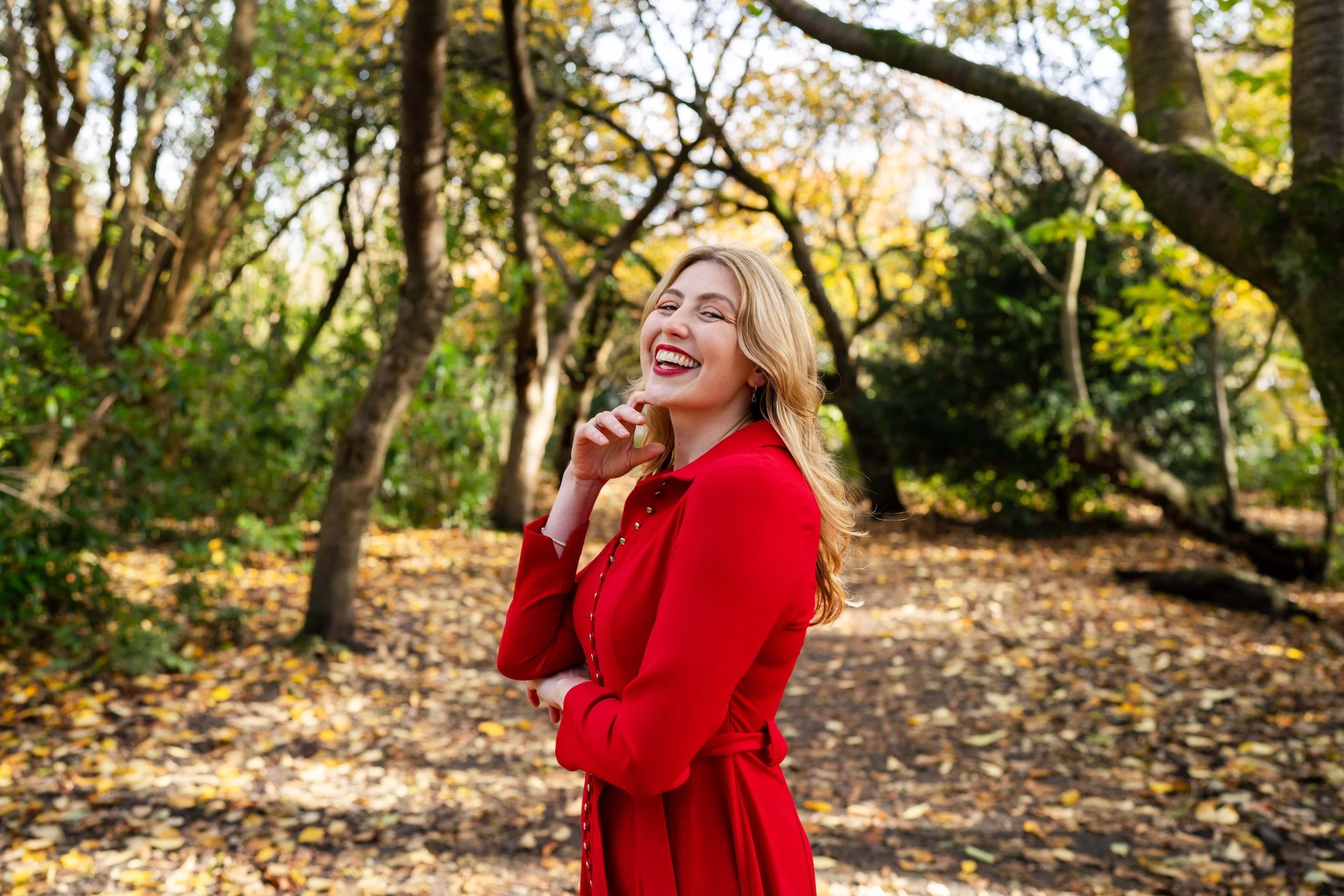 A woman with blonde hair in a red dress smiling in a park with autumn leaves.