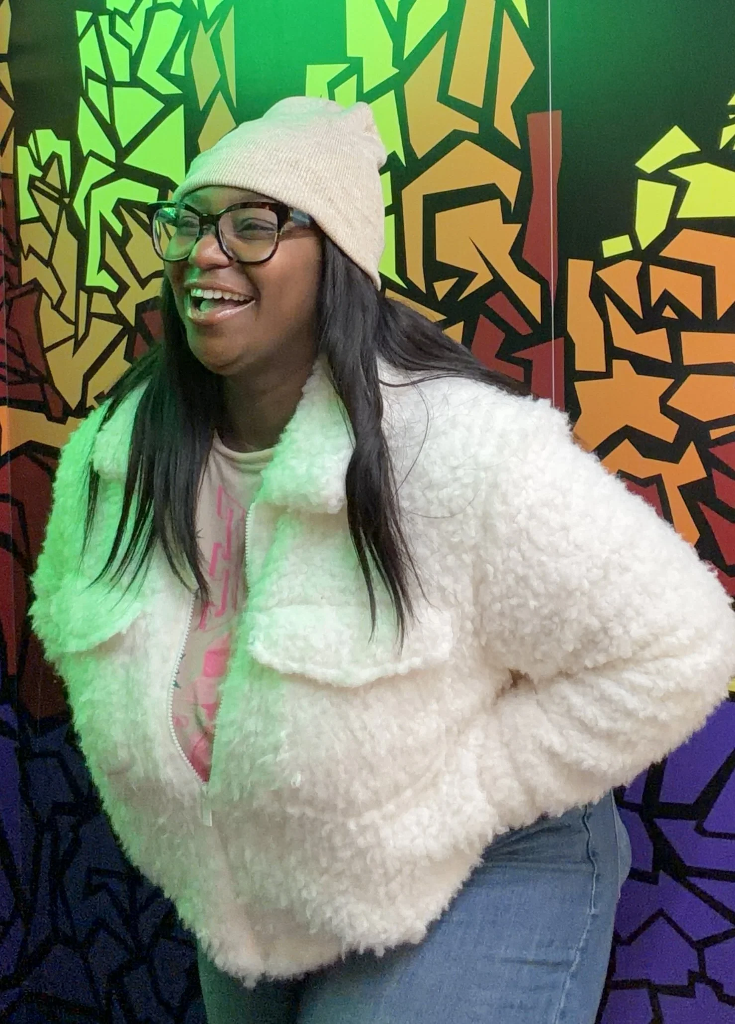 A smiling woman wearing a beige beanie, glasses, and a fluffy white jacket standing in front of a colorful graffiti wall.