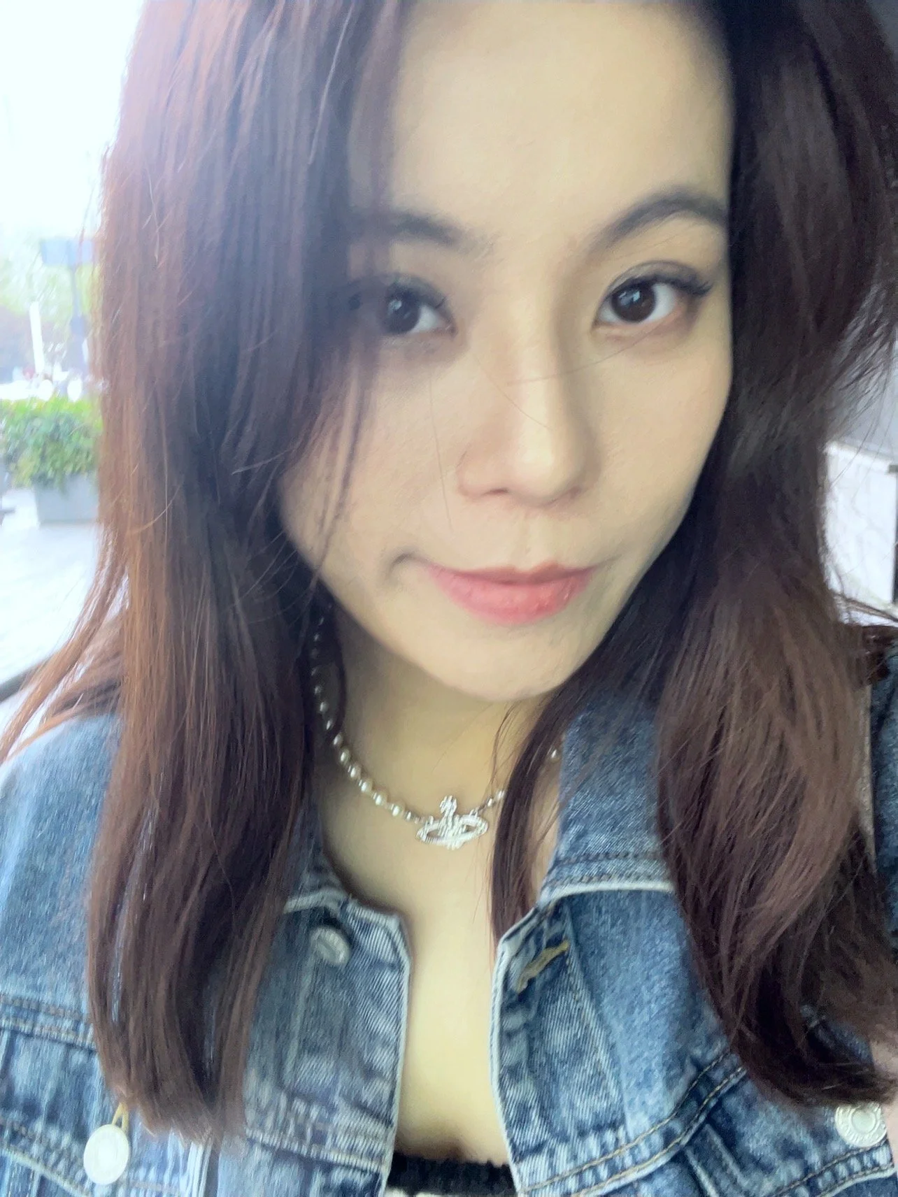 A woman with long brown hair, wearing a denim jacket and a pearl necklace, smiling slightly at the camera.