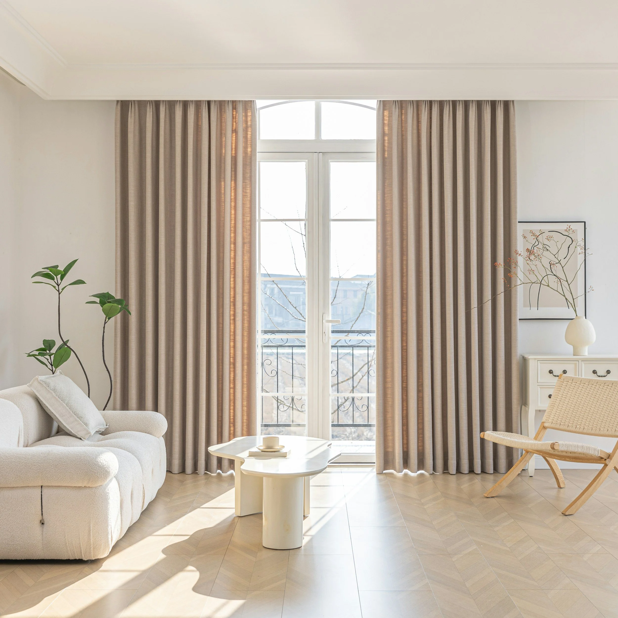 Living room with beige curtains, white sofa, a round coffee table, a wooden chair, a white side table with a vase, and artwork on the wall, illuminated by natural light from a large window.