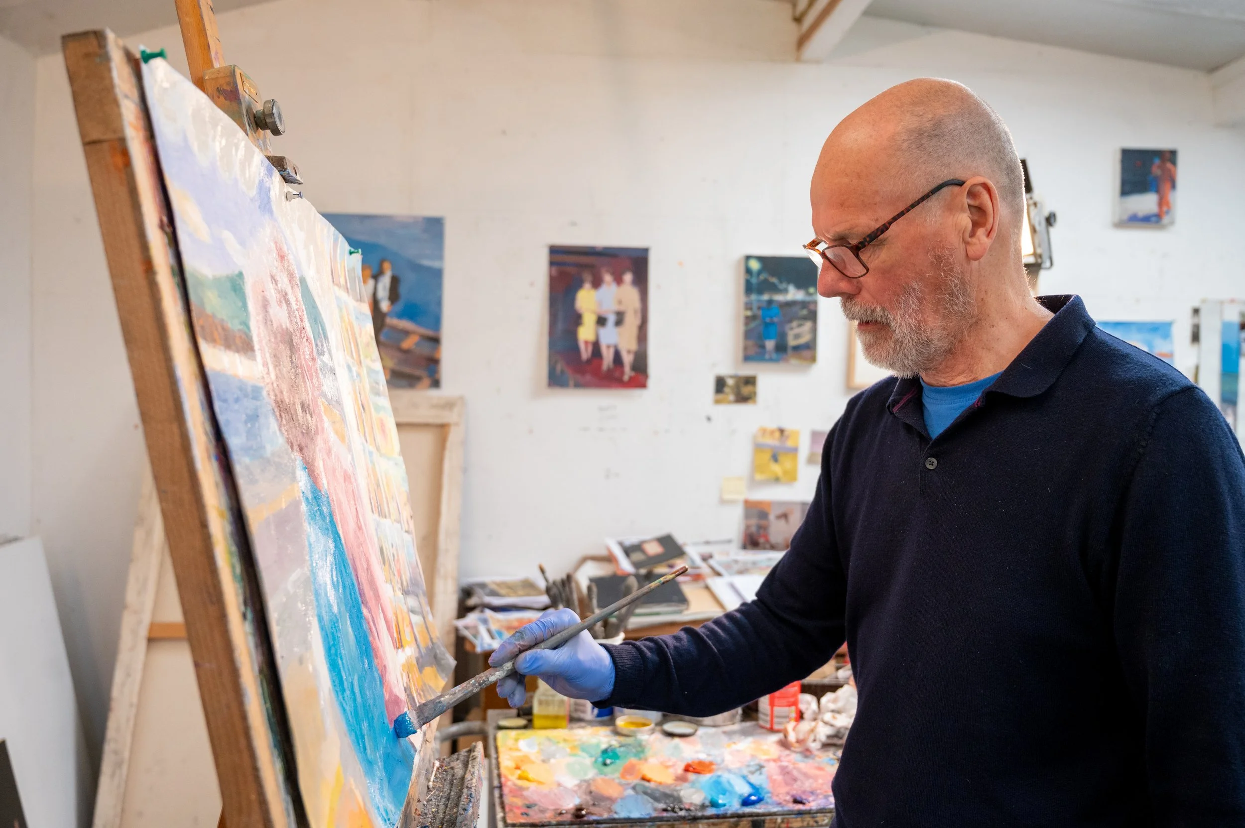 An artist with glasses and a beard is working on a colorful landscape painting on an easel in an art studio. The studio has several paintings hanging on white walls and art supplies on a table nearby. The man is engaging in what matters most to him.