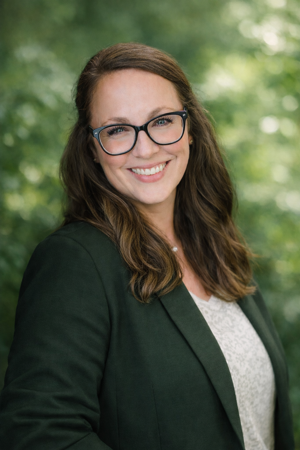 Dr. Allison Ilem, Psychologist specializing in geriatrics. Portrait of a woman with long brown hair, glasses, smiling, wearing a dark blazer and a light-colored blouse, outdoors with blurred green foliage background.