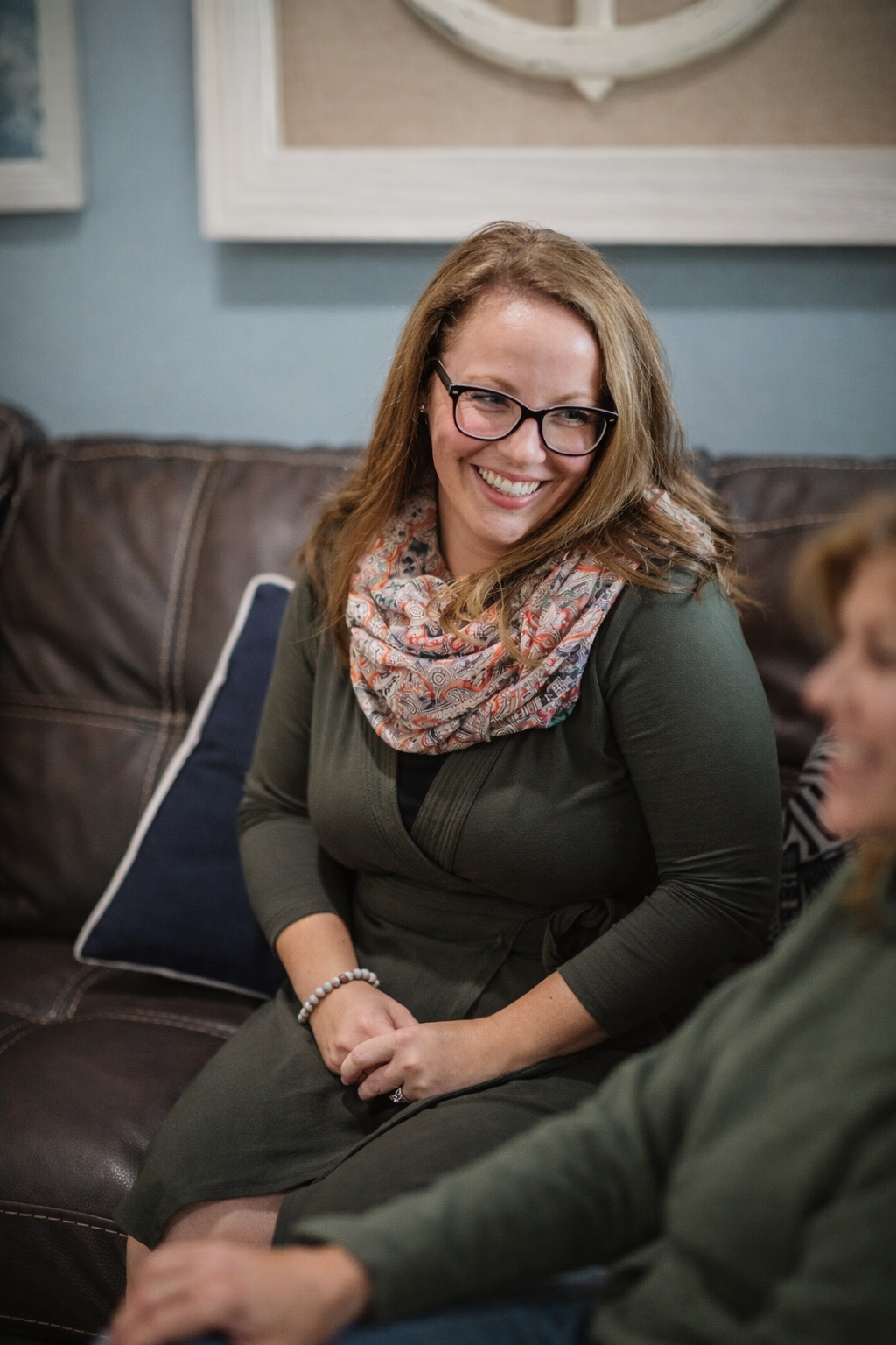 Dr. Allison Ilem, a psychologist specializing in aging and caregiving. She is a woman with glasses and shoulder-length light brown hair smiling and wearing a colorful scarf, sitting on a couch in a living room. She engages with the person next to her