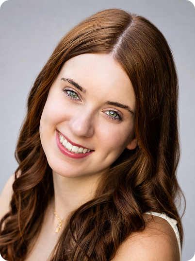 Close-up portrait of a young woman with long, wavy auburn hair, fair skin, and light green eyes, smiling at the camera against a neutral gray background.