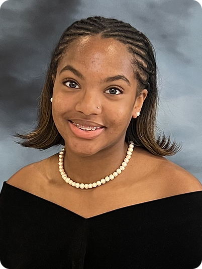 Young woman with medium skin tone, wearing a black off-shoulder top, a pearl necklace, and small earrings, smiling against a neutral gray background.