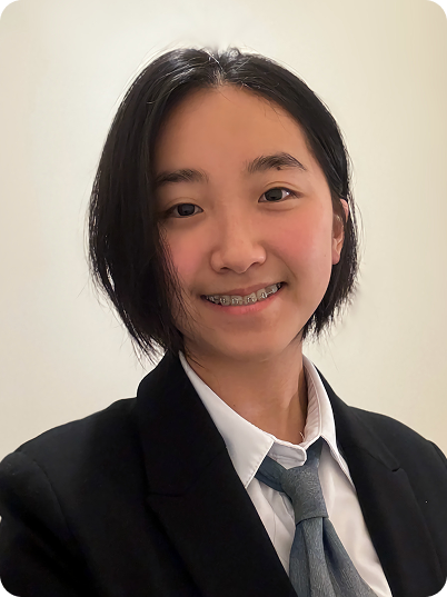 Young girl with shoulder-length black hair, smiling, wearing a school uniform with a blazer, white shirt, and gray tie, against a plain light-colored wall.