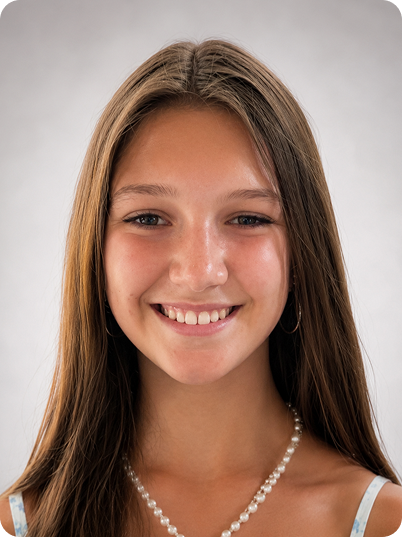 A young woman with long brown hair, smiling, wearing a pearl necklace and a light-colored top, against a plain gray background.