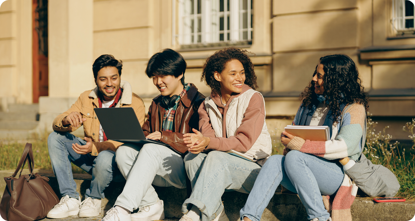 Four young diverse students sitting on a curb outside a building, smiling and talking, with books and a laptop.