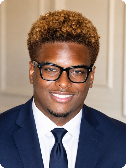 A young man with curly, light brown hair wearing black glasses, a navy blue suit, white shirt, and dark tie, smiling indoors.