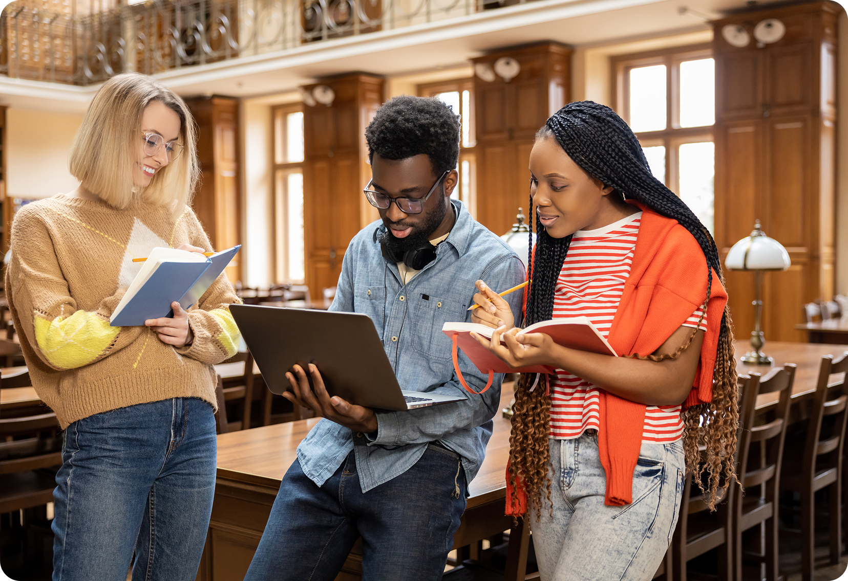 Three diverse students studying together in a library, discussing notes and using a laptop.