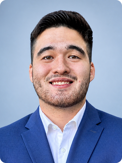 Portrait of a young man with dark hair, facial hair, wearing a blue suit and white shirt, smiling, against a light blue background.