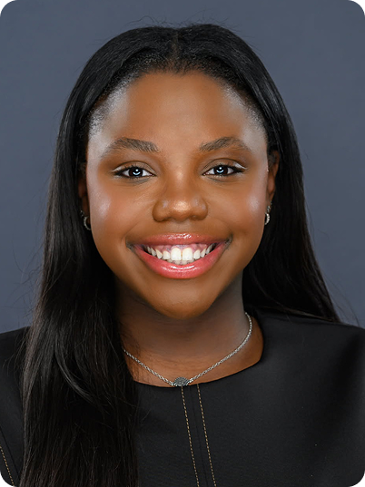 Close-up of a smiling young woman with long black hair, wearing a black top, a silver necklace, and earrings against a dark gray background.