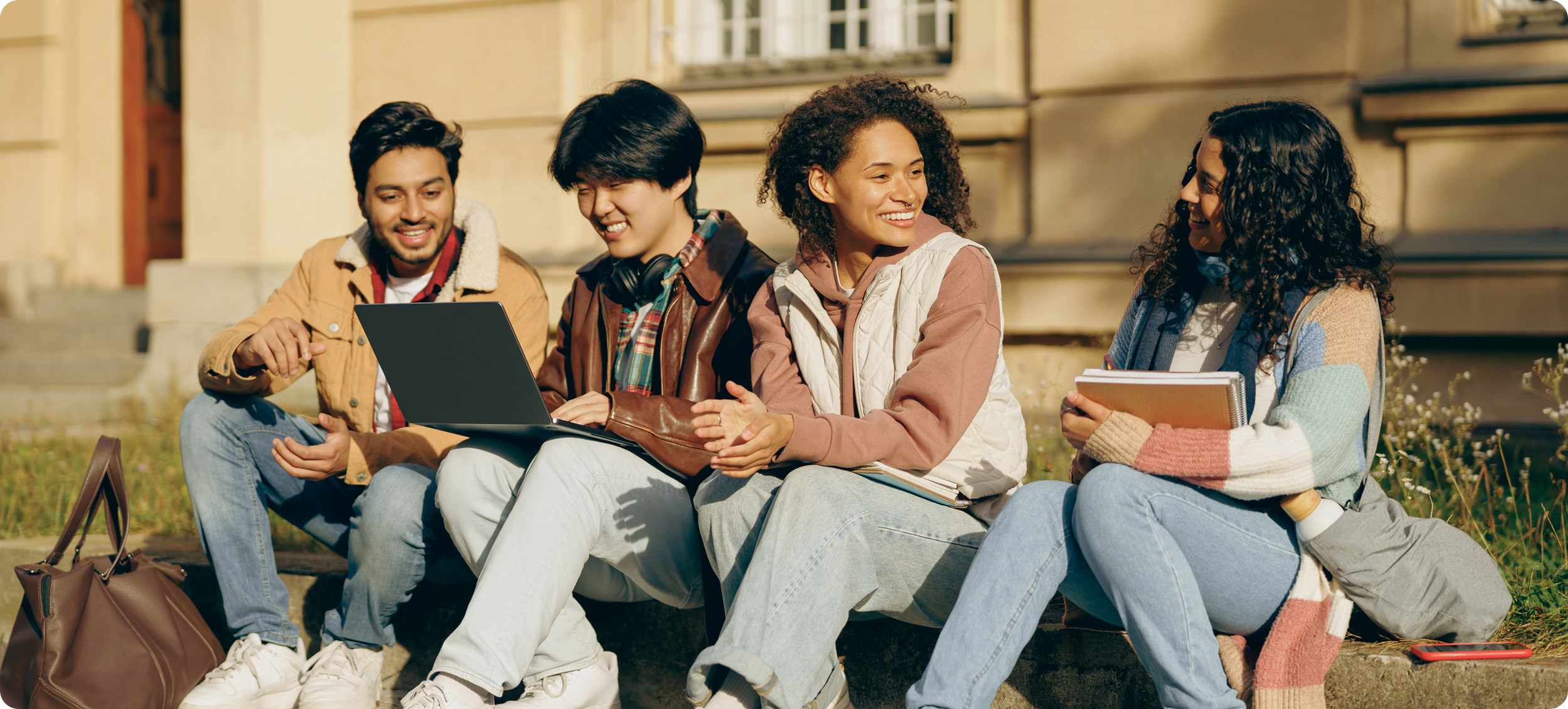 Four diverse young adults sitting on a stone bench outdoors, engaging in conversation and using a laptop and notebooks, with a historic building in the background.