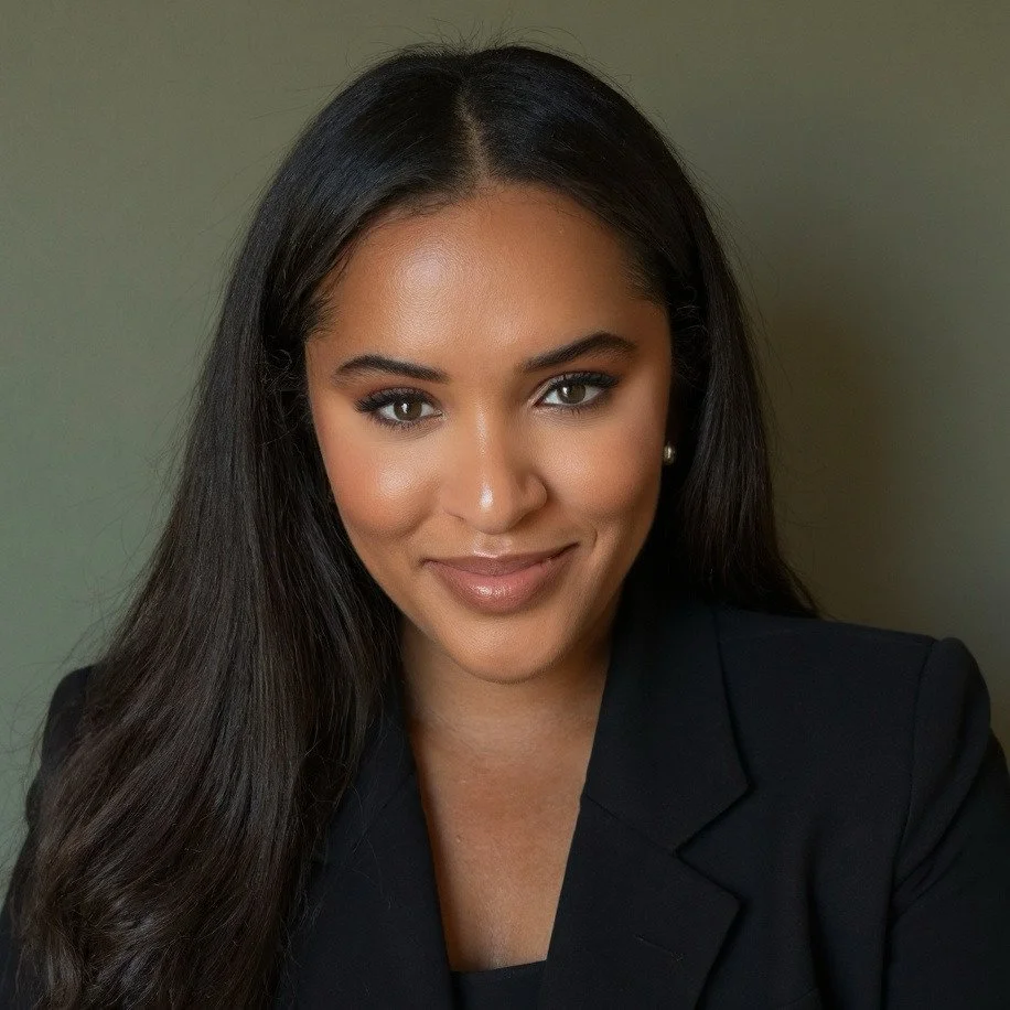 A woman with long dark hair, makeup, wearing earrings and a black blazer, smiling at the camera against a plain background.