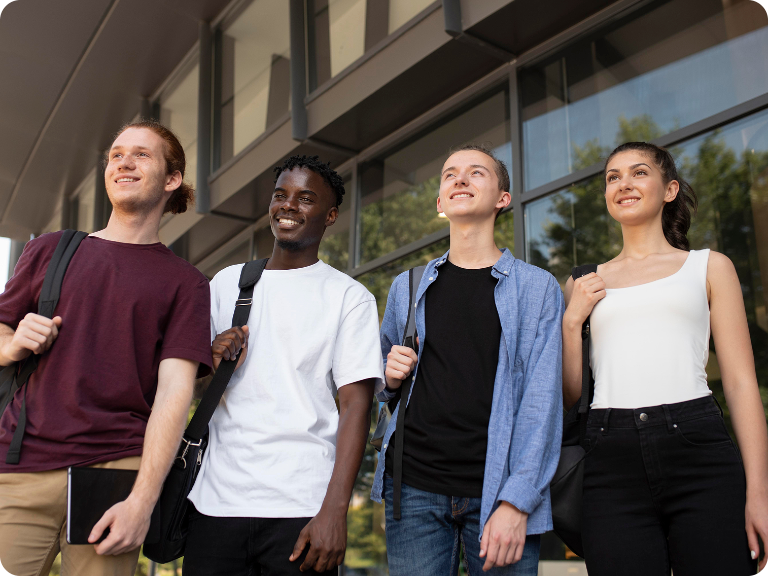 Four young adults standing outside a modern building, smiling and looking upward, carrying backpacks.