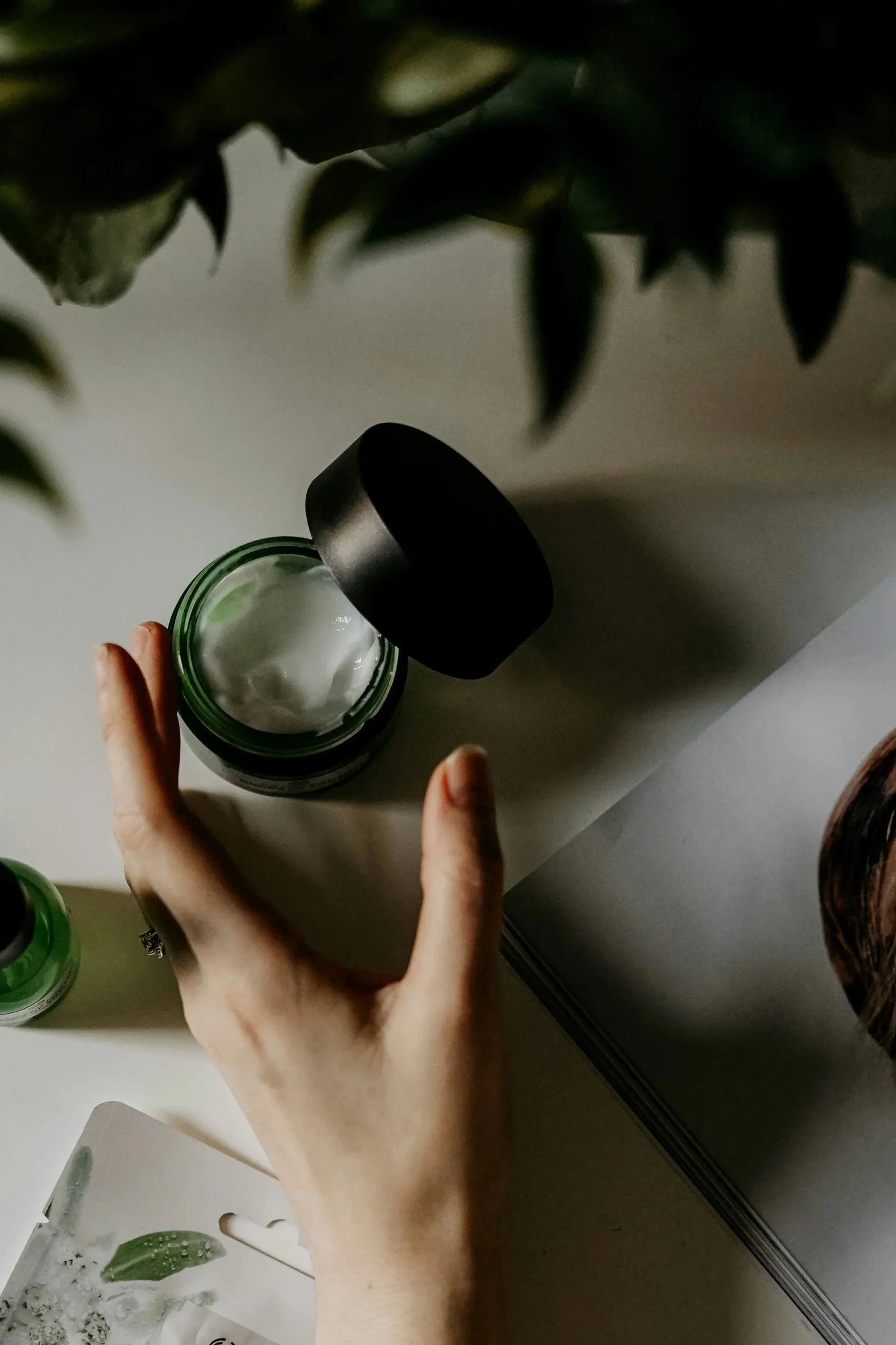 Une main tenant un pot ouvert contenant une crème blanche, avec un couvercle noir à côté, posé sur un surface blanche. Des plantes vertes sont visibles en haut de l'image.