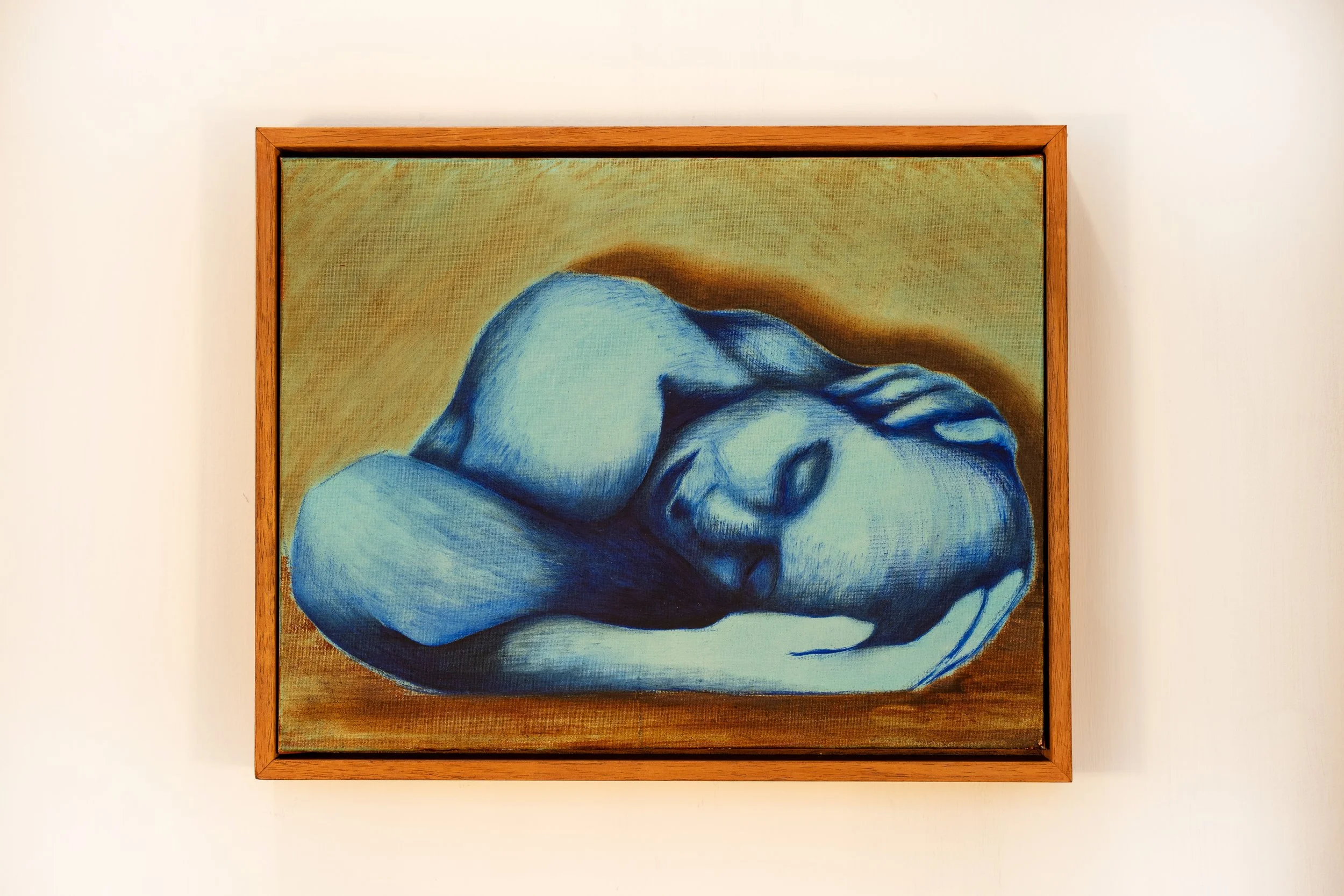 A painting of a woman with blue tones, lying curled up with her eyes closed, framed in wood and hanging on a white wall.