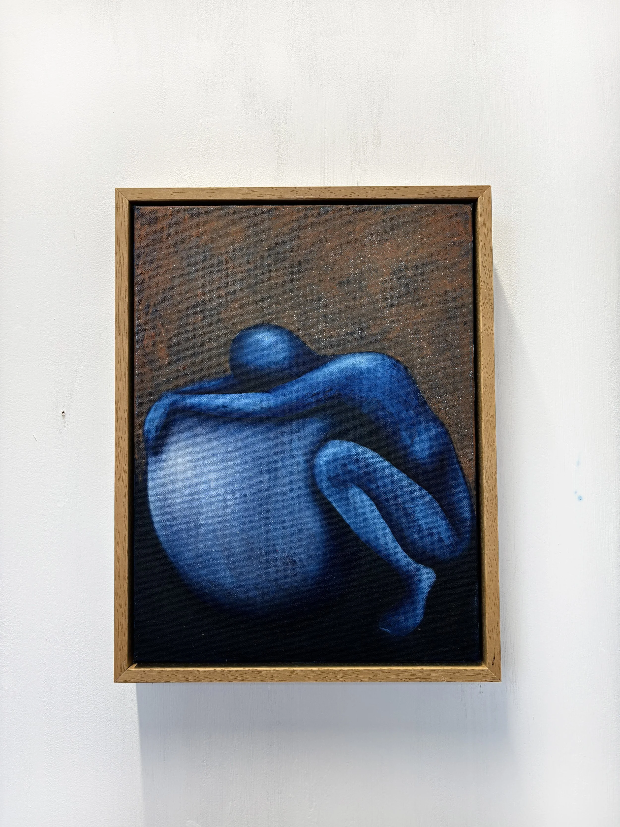 A painting of a blue humanoid figure curled into a fetal position, with a brown background, framed in wood, hanging on a white wall.