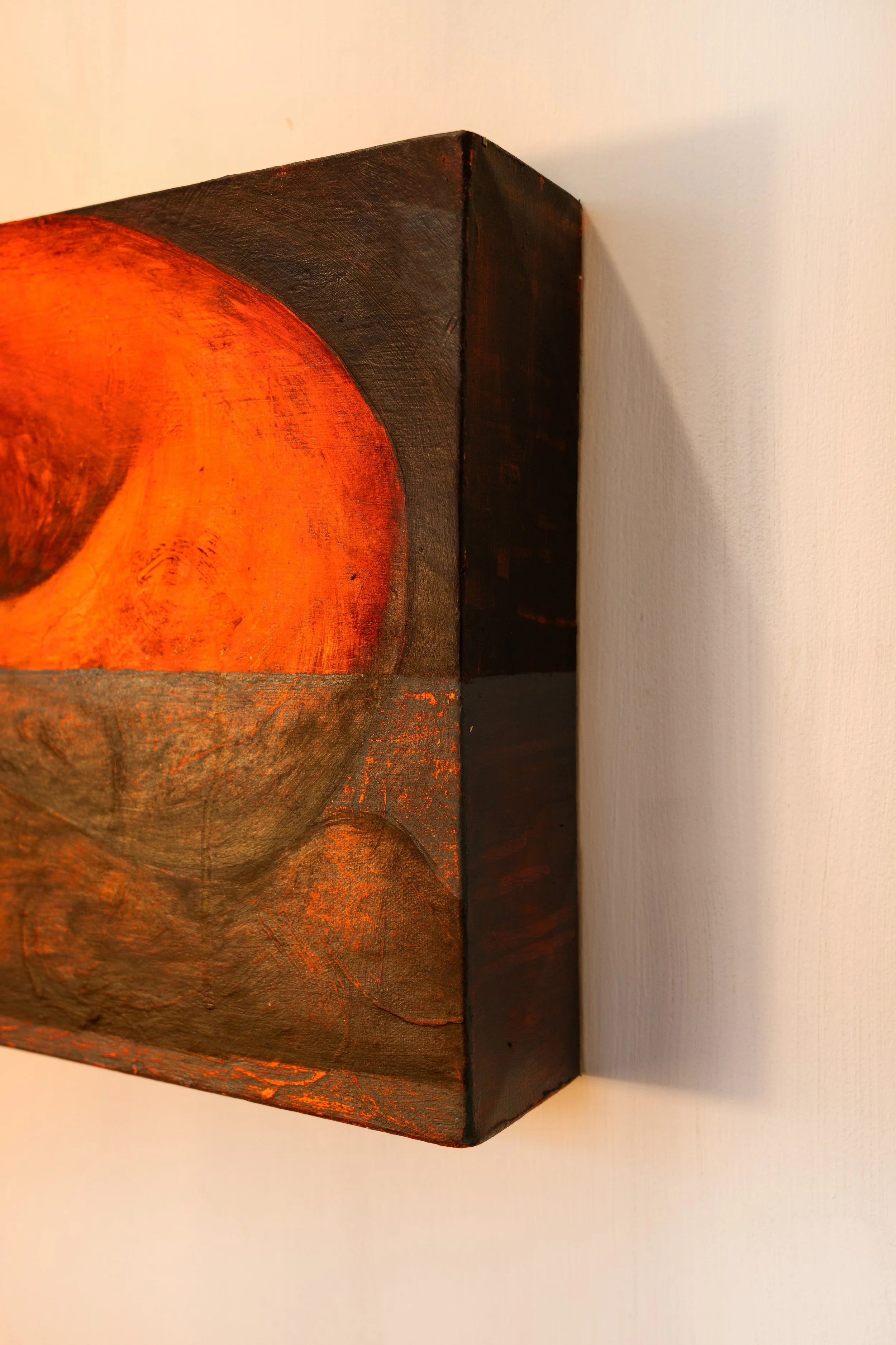 Close-up of a textured, painted wooden art piece featuring an abstract circular design with warm orange and brown tones, mounted on a white wall.